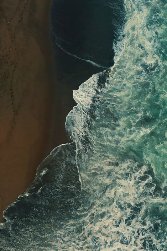 Aerial view of ocean waves with white foam crashing against a dark sandy beach, showing deep teal water and brown sand in a high-contrast composition.