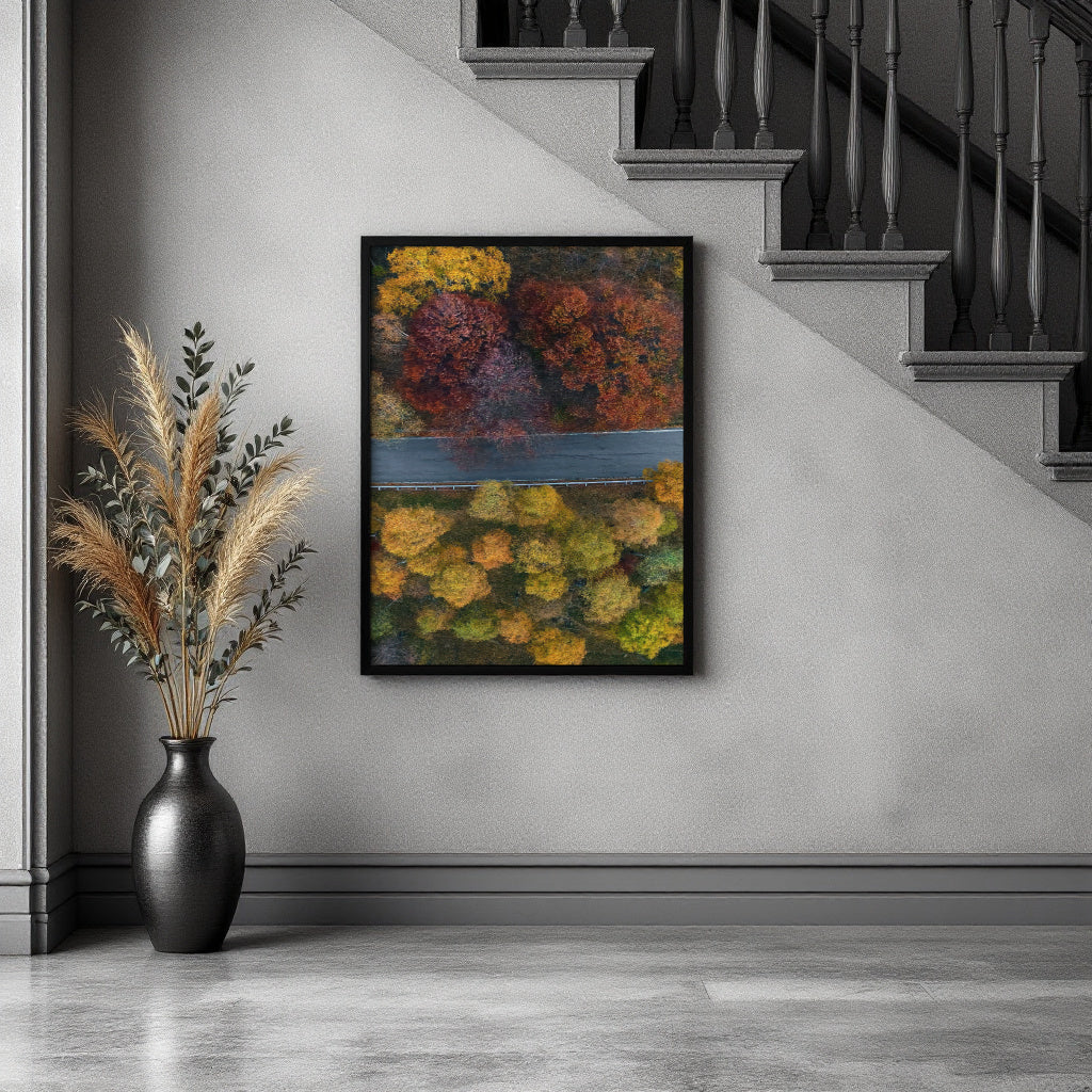 A framed abstract landscape painting featuring vibrant autumn foliage in yellow, orange, red, and green tones, hanging on a gray wall in a modern interior setting next to a black vase with dried grasses and below a staircase.