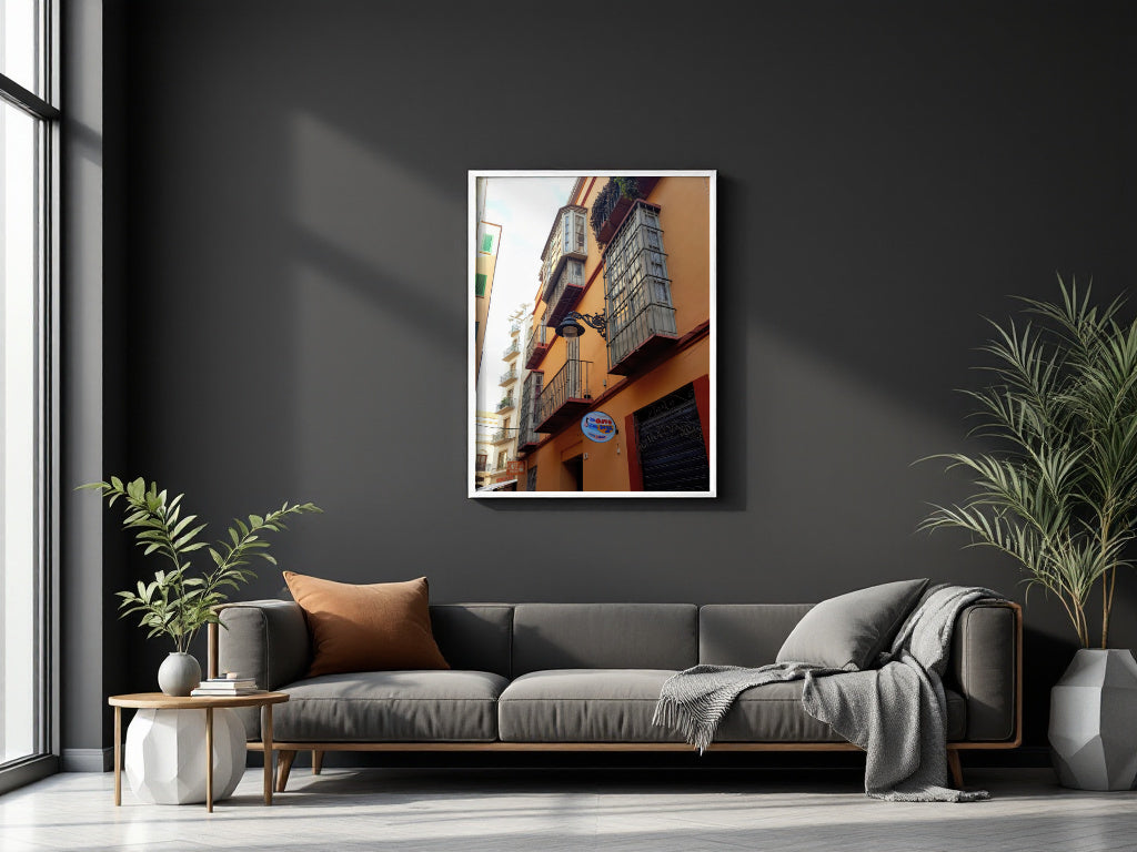 A framed vertical art print of a warm-toned building facade hangs on a dark charcoal wall above a modern gray sofa in a styled living room setting.