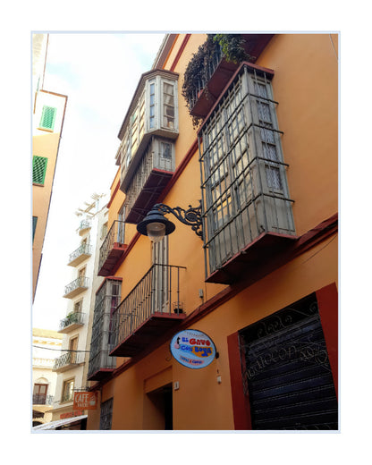An upward-looking photograph of a warm orange apartment building facade with multiple balconies, metal shutters, a hanging street lamp, and a round blue-and-white shop sign reading 'el guapo'.