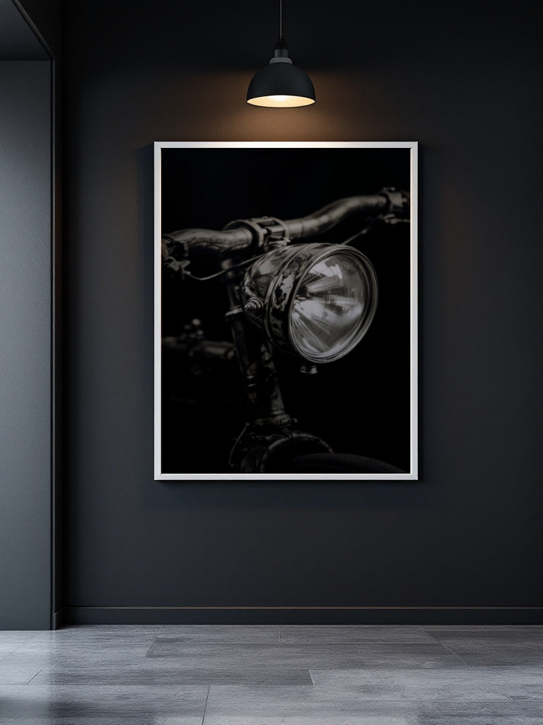 A framed black and white art print of a vintage bicycle headlight hanging on a dark charcoal wall under a hanging lamp.