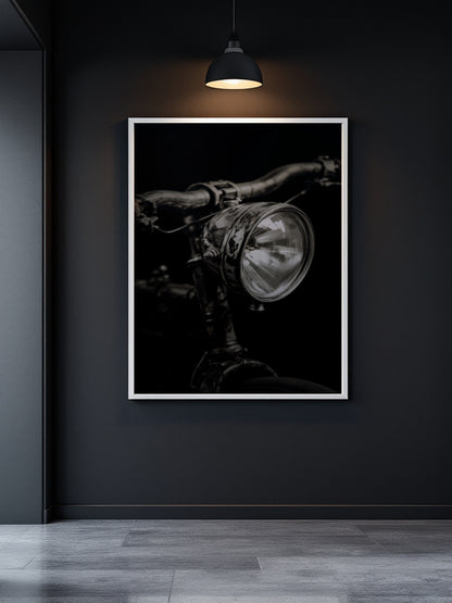 A framed black and white art print of a vintage bicycle headlight hanging on a dark charcoal wall under a hanging lamp.