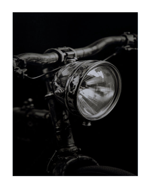 A black and white close-up photograph of a vintage bicycle headlight mounted on dark metal handlebars against a black background.