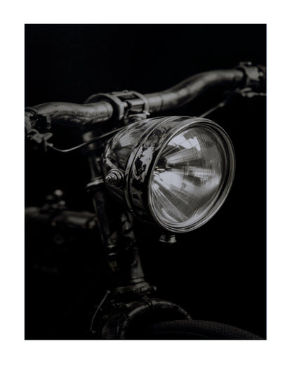 A black and white close-up photograph of a vintage bicycle headlight mounted on dark metal handlebars against a black background.