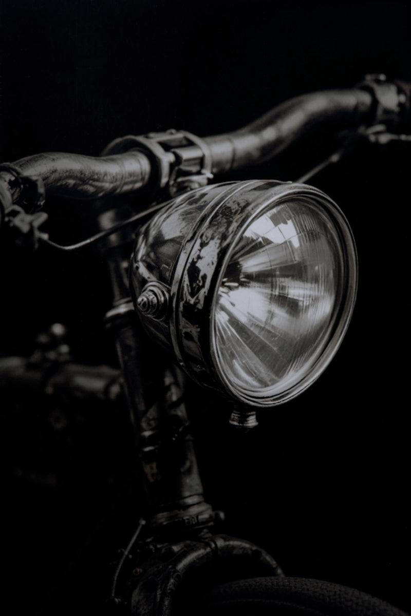 A close-up, dramatic black and white photograph of a vintage bicycle headlight mounted on dark handlebars against a solid black background.
