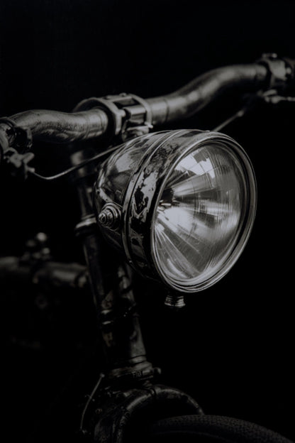 A close-up, dramatic black and white photograph of a vintage bicycle headlight mounted on dark handlebars against a solid black background.