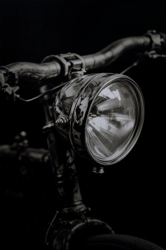 A close-up, dramatic black and white photograph of a vintage bicycle headlight mounted on dark handlebars against a solid black background.