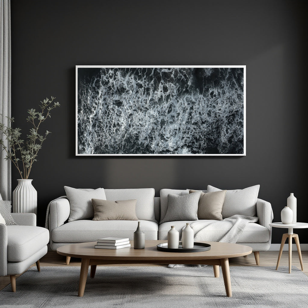 A large framed black and white abstract wave or foam print hangs on a dark charcoal wall above a light gray sofa in a modern living room setting.