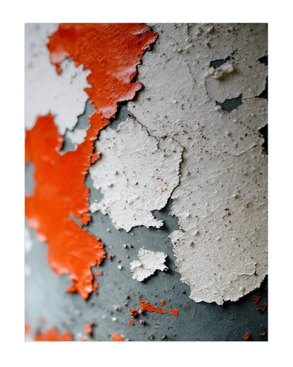 Abstract close-up of peeling paint with bright orange-red layers over a dark gray surface and large areas of off-white chipped paint.