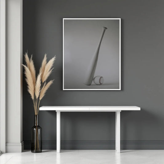 A framed black and white art print depicting a minimalist vase and ball, displayed on a dark gray wall above a white console table with a vase of pampas grass.