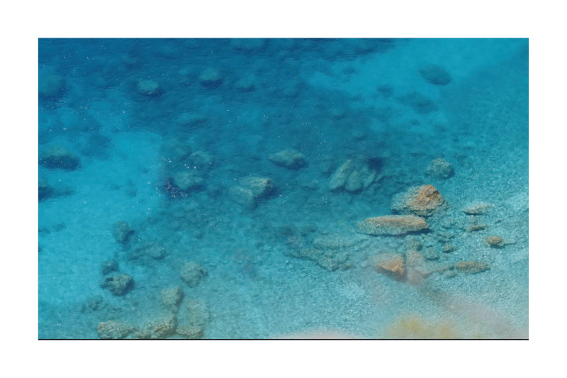 Underwater photograph of a clear blue ocean floor with scattered rocks and sandy seabed visible through shallow turquoise water.