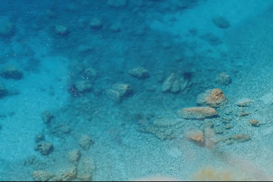 Underwater clear blue ocean water showing a sandy seabed with scattered rocks and coral formations.
