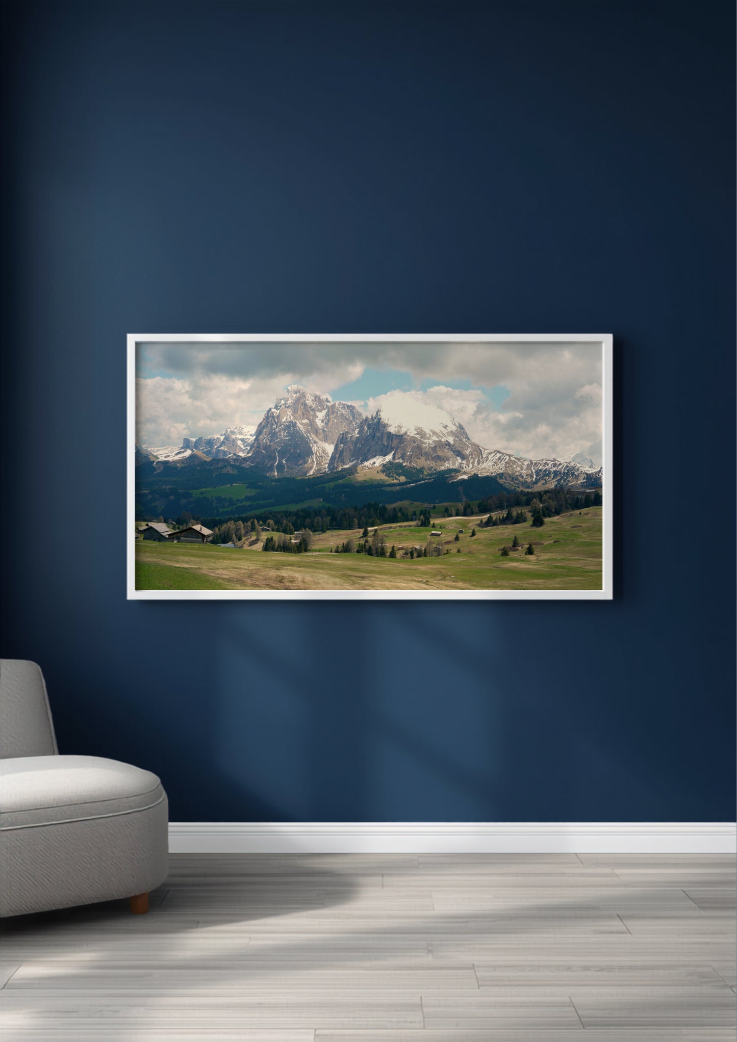A framed landscape art print of snow-capped mountains and green valleys, displayed on a dark navy blue wall above a light gray floor, with a partial view of a gray armchair on the left.