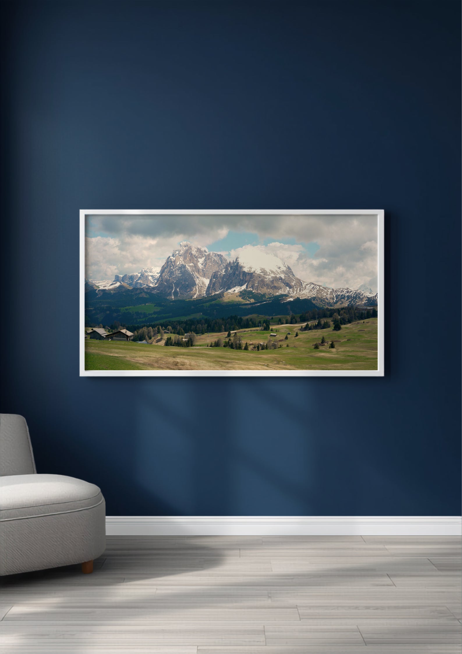 A framed landscape art print of snow-capped mountains and green valleys, displayed on a dark navy blue wall above a light gray floor, with a partial view of a gray armchair on the left.