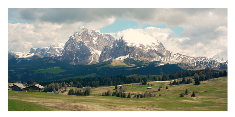 A scenic landscape photograph of a rugged snow-capped mountain range under a cloudy sky, with green hills, forests, and small buildings in the foreground.