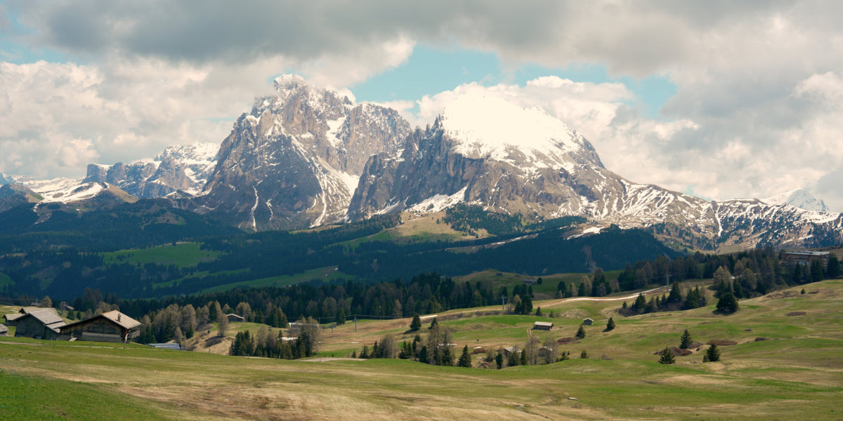 A scenic mountain landscape with snow-capped peaks, green valleys, scattered trees, and a few small buildings under a cloudy sky.