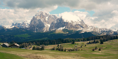 A scenic mountain landscape with snow-capped peaks, green valleys, scattered trees, and a few small buildings under a cloudy sky.