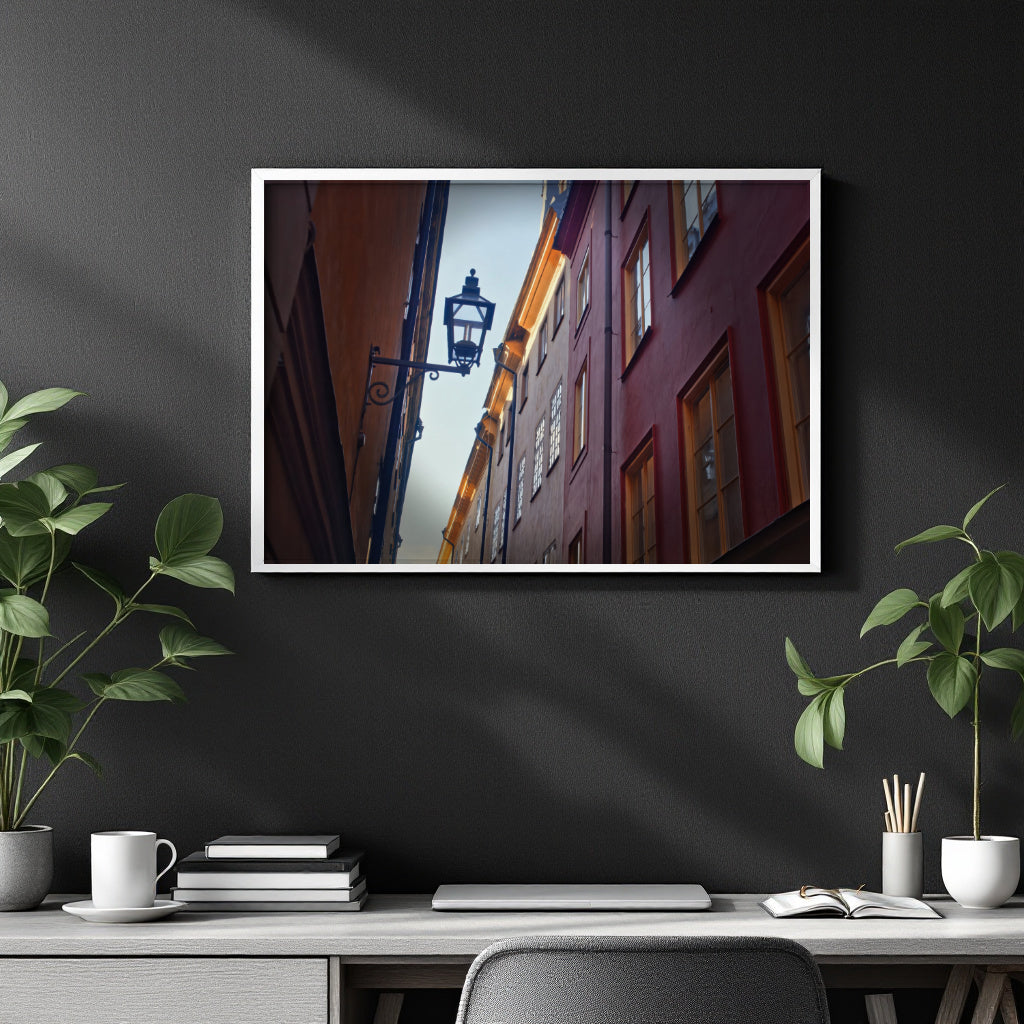 A framed vertical art print depicting a narrow European street with red and orange buildings, hanging on a dark gray wall above a styled desk with plants and books.