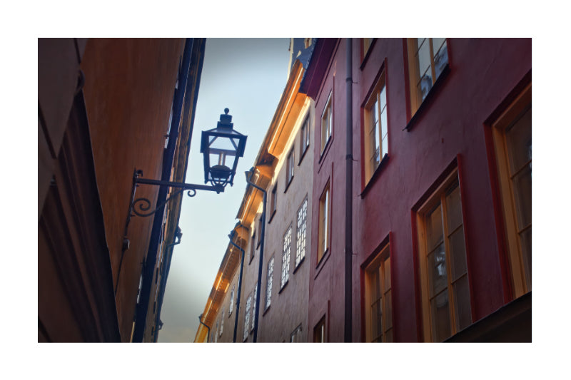 A vertical photographic print of a narrow European street with warm red and brown buildings, glowing windows, and a black metal street lamp on the left, shown with a white border.