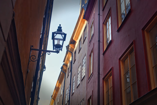 A narrow European street with tall buildings in reddish-brown and orange tones, featuring a black metal wall lamp and warm window light against a pale sky.