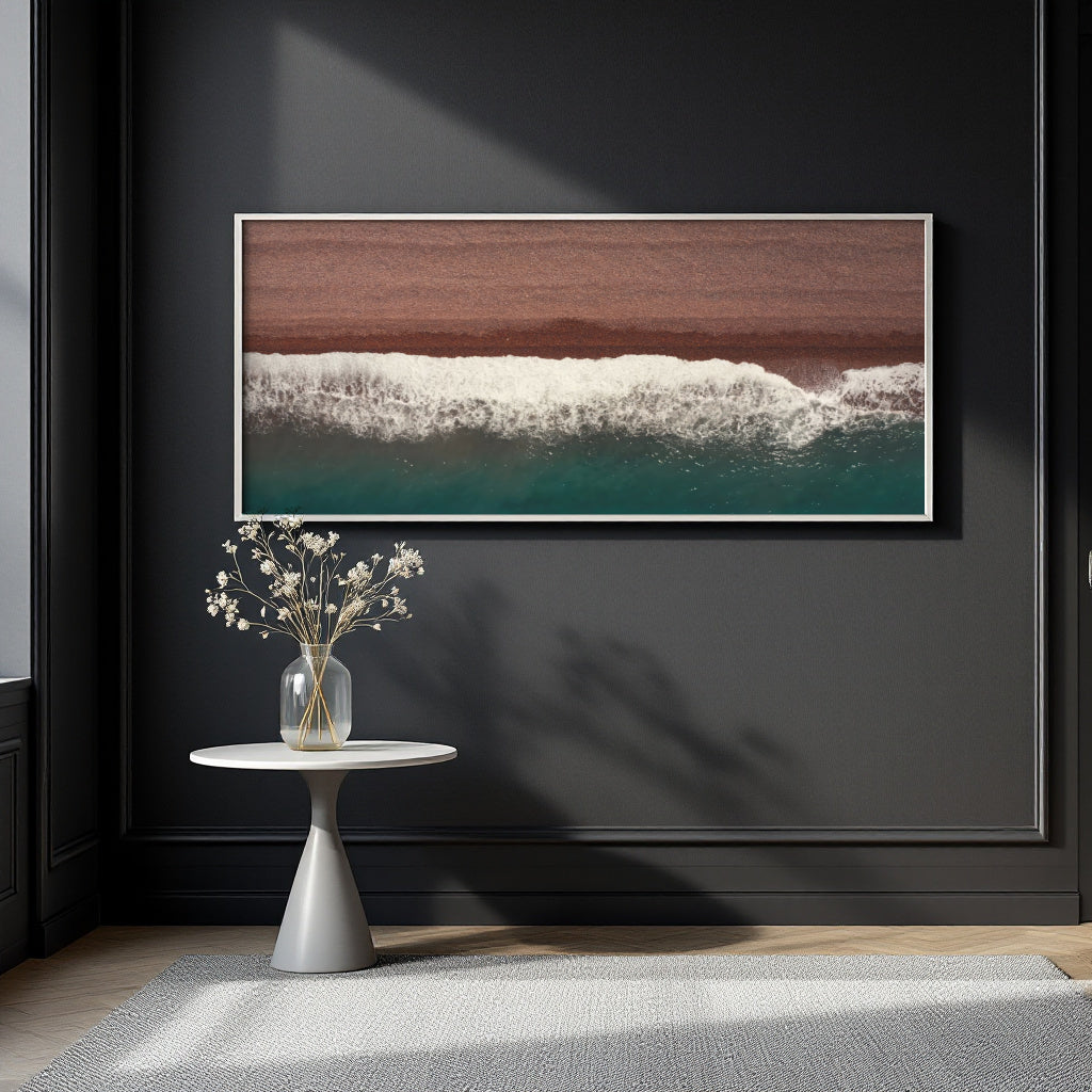 A framed horizontal wall art print showing a beach shoreline with a brown sand section, white surf, and teal water, displayed on a dark charcoal wall above a small white side table with a vase of flowers.