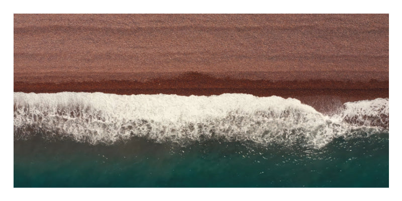 Aerial view of ocean waves with white foam rolling onto a reddish-brown shoreline under a teal sea.
