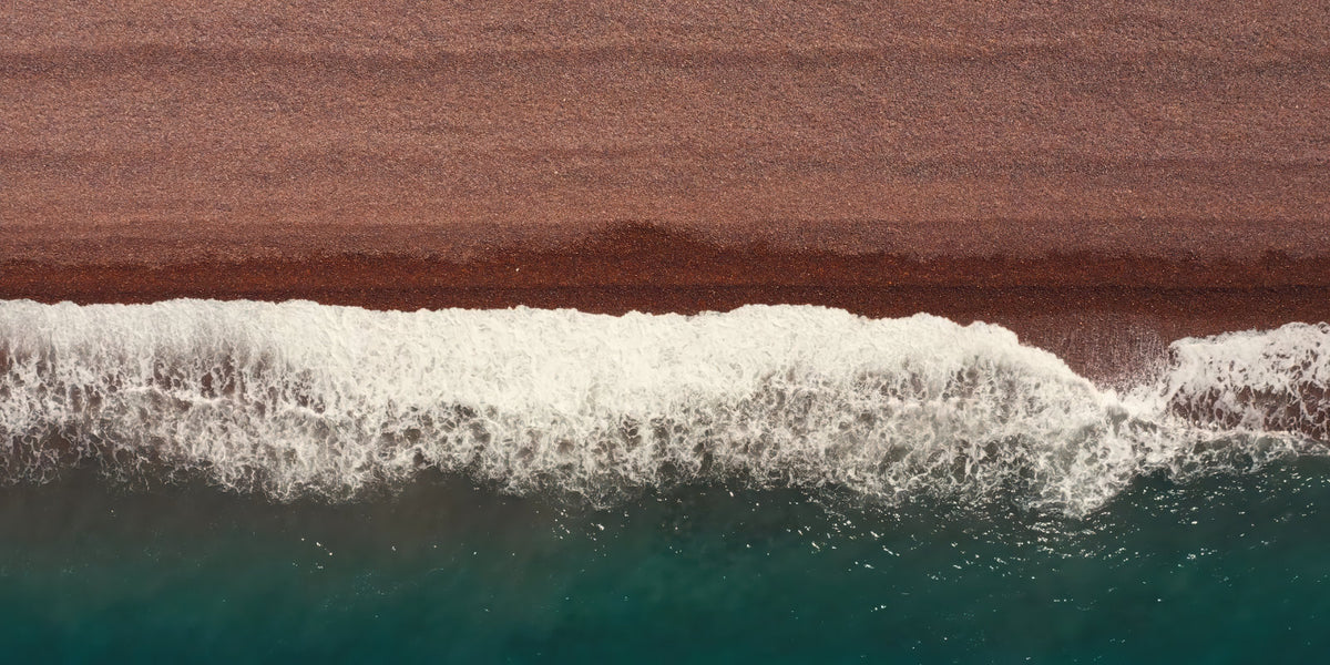 Aerial view of ocean waves with white foam rolling into deep teal water, bordered by a wide brown sandy shoreline.