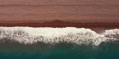 Aerial view of ocean waves with white foam rolling into deep teal water, bordered by a wide brown sandy shoreline.