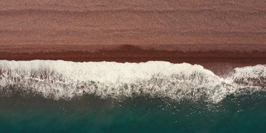 Aerial view of ocean waves with white foam rolling into deep teal water, bordered by a wide brown sandy shoreline.