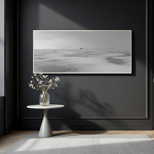 A black and white panoramic landscape art print with a thin white frame, displayed on a dark charcoal wall above a small white side table with a glass vase of dried flowers.