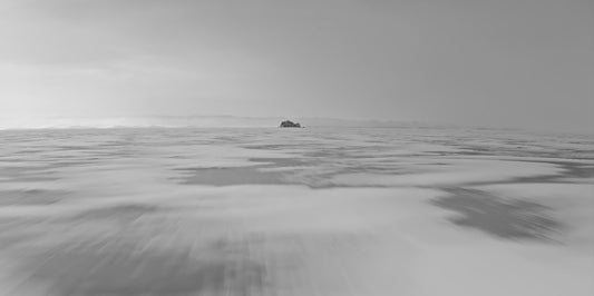 A minimalist black and white landscape photograph featuring a small, dark rock formation situated in the center of a vast, misty sea or desert dune field under a cloudy sky.