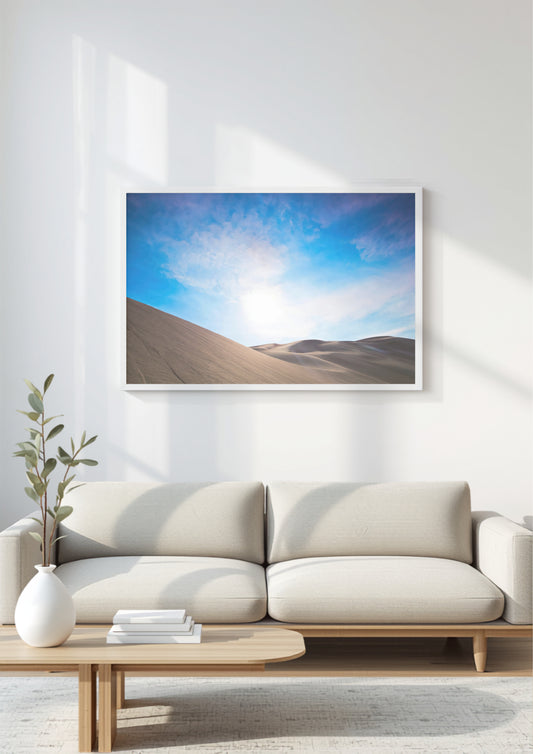 A framed landscape art print of desert sand dunes under a bright blue sky, displayed on a white wall above a modern beige sofa in a minimalist living room setting.