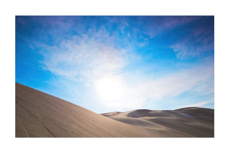 A scenic landscape photograph of desert sand dunes under a bright blue sky with wispy white clouds and a glowing sun near the horizon.