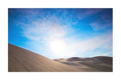 A scenic landscape photograph of desert sand dunes under a bright blue sky with wispy white clouds and a glowing sun near the horizon.