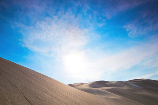 A scenic landscape photograph of rolling sand dunes under a bright blue sky with wispy white clouds and a glowing sun near the horizon.