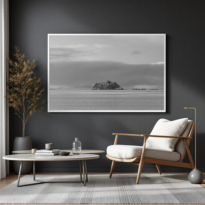 A large black and white framed landscape art print of a rocky island on the sea, displayed on a dark gray wall in a modern living room setting with a wooden armchair, round coffee table, and a potted plant.
