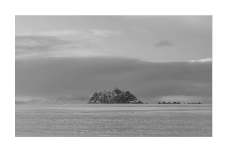 A black and white landscape photograph of a small rocky island rising from calm water under a cloudy sky.