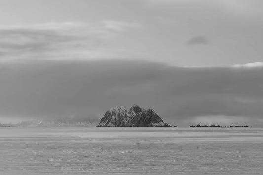 Black and white landscape photograph of a small rocky island rising from calm water under a cloudy sky.