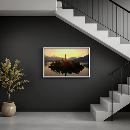 A framed landscape art print of Bled Island in Slovenia, showing a church on an island reflected in the water at sunset, displayed on a dark gray wall in a modern interior setting with a staircase and a potted plant.