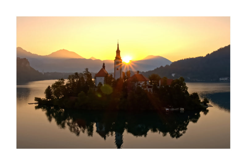 A scenic landscape print of Lake Bled in Slovenia at sunset, featuring the Church of Our Lady of Mount Carmel on a small island with its reflection in the calm water, surrounded by mountains under a golden sky.