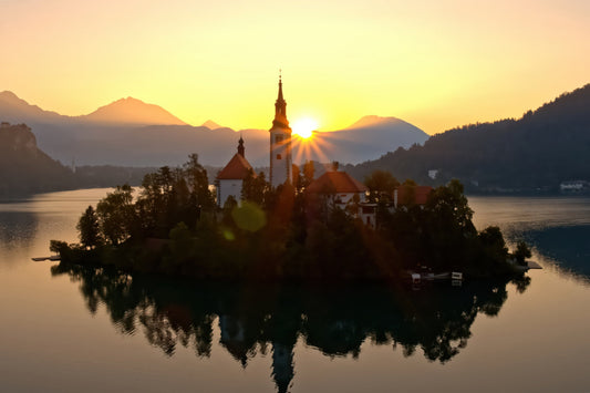 A scenic landscape photograph of Bled Lake in Slovenia at sunset, featuring the Church of Our Lady of Bled on a small island with its reflection in the calm water, surrounded by mountains and a golden sky.