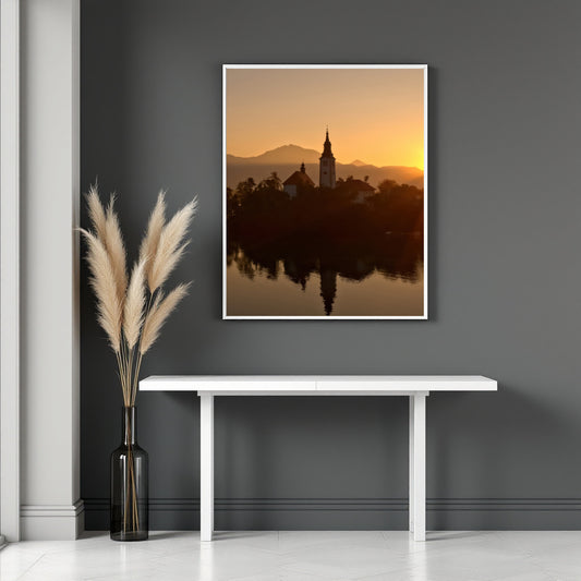 A framed vertical wall art print showing a sunset landscape with a church tower reflected in water, displayed above a white console table against a dark gray wall. A tall vase with pampas grass stands to the left.