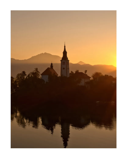 A scenic landscape photograph of a church spire and buildings on a small island, silhouetted against a warm orange sunset over distant mountains, with the reflection visible in the water.