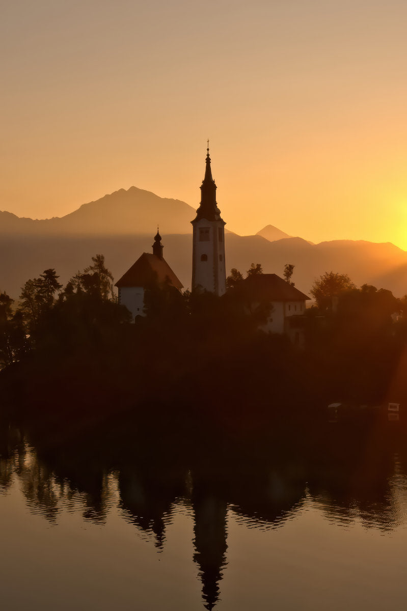 A scenic landscape photograph of a church tower and buildings on a small island in a lake, silhouetted against a warm golden sunset with mountain ranges in the background and reflections in the water.