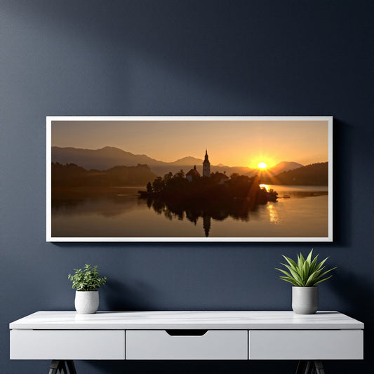 A framed panoramic landscape print of Lake Bled, Slovenia, featuring a sunset over an island with a church spire, displayed on a dark blue wall above a white console table with two small potted plants.
