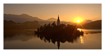 A scenic landscape print of a lake at sunset with a small island, a church spire, and mountains in the background, shown in warm golden tones.