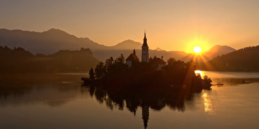 A scenic landscape photograph of a small island church on a lake at sunset, with mountains in the background and the sun low on the horizon.