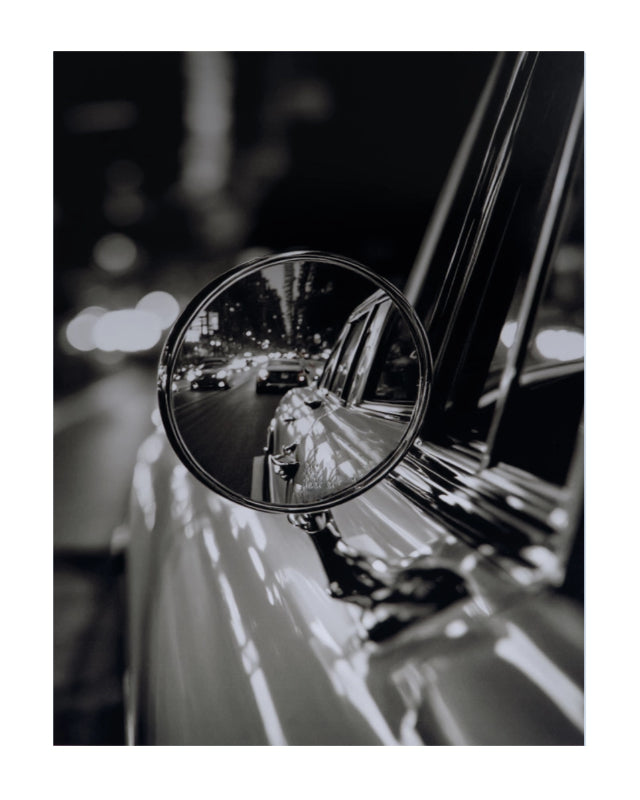 Black and white photograph of a car side mirror reflecting a city street at night, presented as an art print with a white border.
