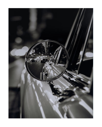 Black and white photograph of a car side mirror reflecting a city street at night, presented as an art print with a white border.