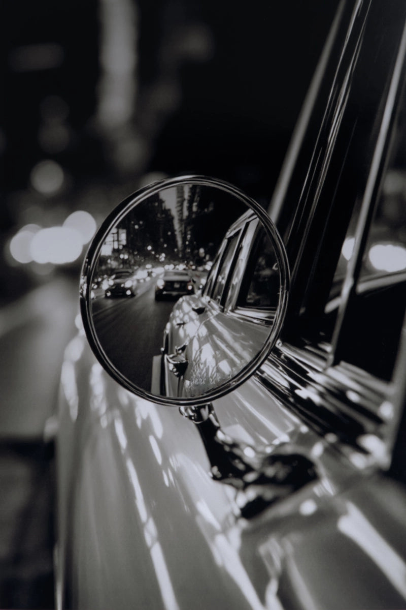 A black and white close-up photograph of a classic car's side-view mirror mounted on the door, reflecting a blurred city street at night.
